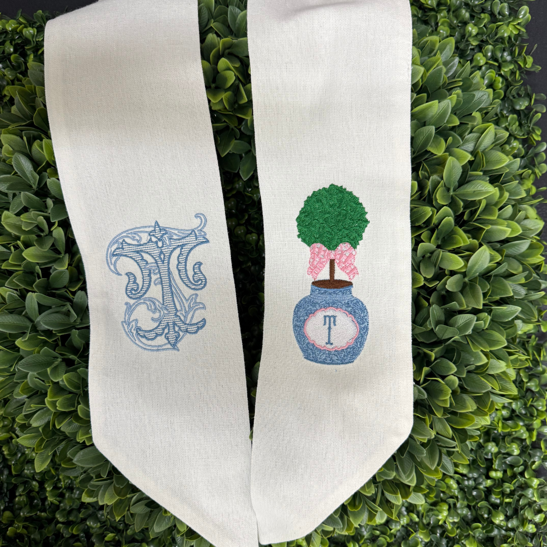 A white sash with embroidered initials and an embroidered topiary with a pink bow, the letter "T" on a blue and white planter